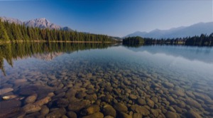 Calm clear lake in the canadian rockies with the rocks on the lake bed in forground treeline and mountains in background